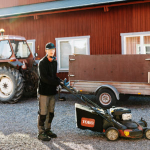 En elev med en gräsklippare. I bakgrunden en traktor med släp framför en röd ladugård.